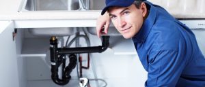 An emergency plumber in Atlanta wearing a blue uniform works under a kitchen sink, looking up while holding a wrench.