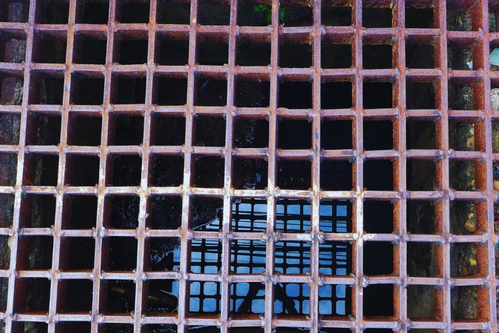 Rusty metal grate covering a square sewage drain with water visible beneath.