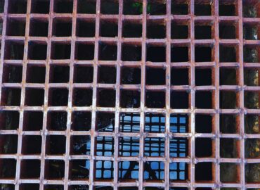 Rusty metal grate covering a square sewage drain with water visible beneath.