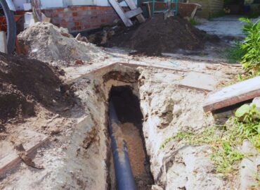 A partially dug-up yard shows a trench containing a visible underground pipe. Dirt piles are placed nearby, and various tools are scattered around the site—a reminder of how crucial homeowners insurance can be for unexpected issues like sewer backup.