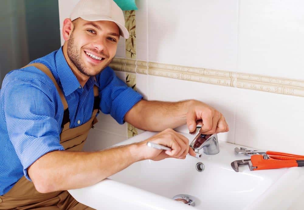 A person in work attire adjusts a faucet with a wrench in a bathroom setting, smiling at the camera, showcasing their skills in burst pipe repair.